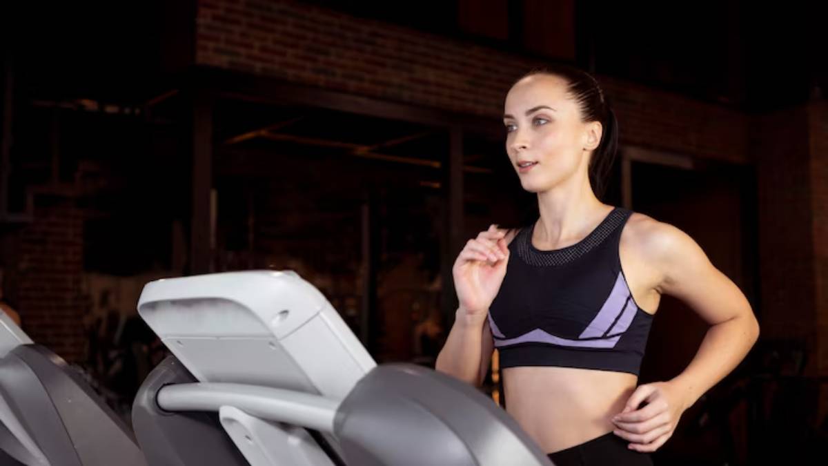 A person in a black athletic crop top exercises on a treadmill in a brightly lit gym with exposed brick walls.