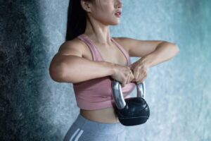 A person in a pink sports top lifts a kettlebell, showcasing strength and focus against a soft blue-gray background.