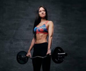 A muscular individual stands confidently holding a barbell, showcasing toned abs and wearing a colorful sports top and black leggings.