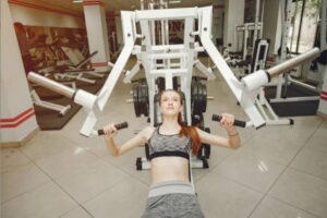 A person exercises on a weight lifting machine in a bright gym, surrounded by various fitness equipment and mirrors.