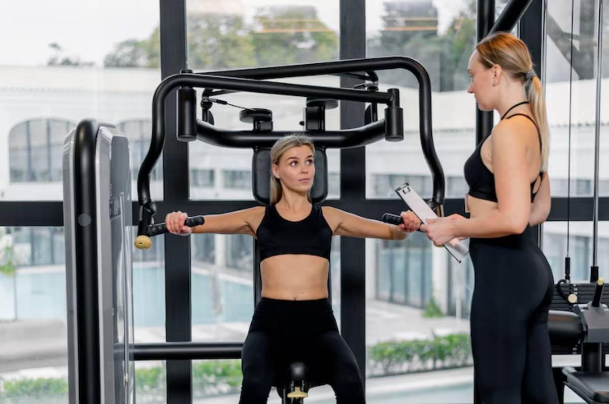 A person exercises on a machine in a bright gym, while a trainer provides guidance, holding a clipboard. Large windows offer a view of greenery.