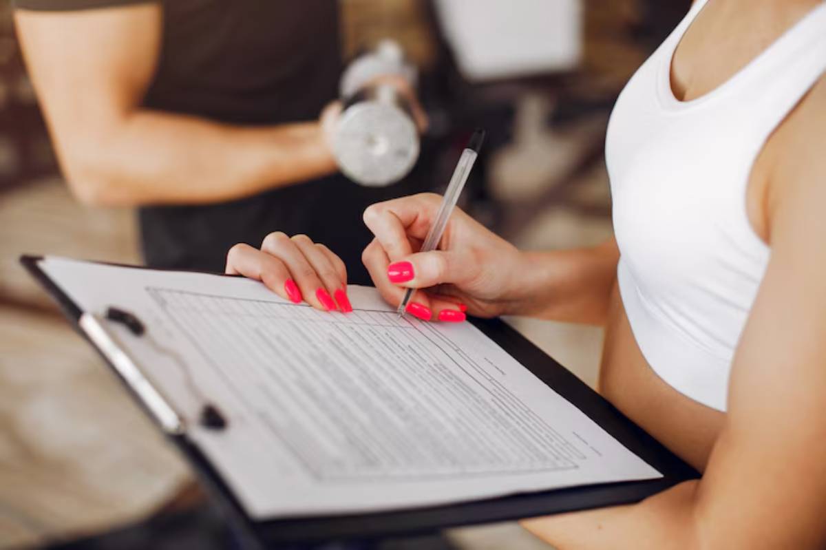 A woman with bright pink nails writes on a clipboard in a gym, while a man with a dumbbell stands in the background.