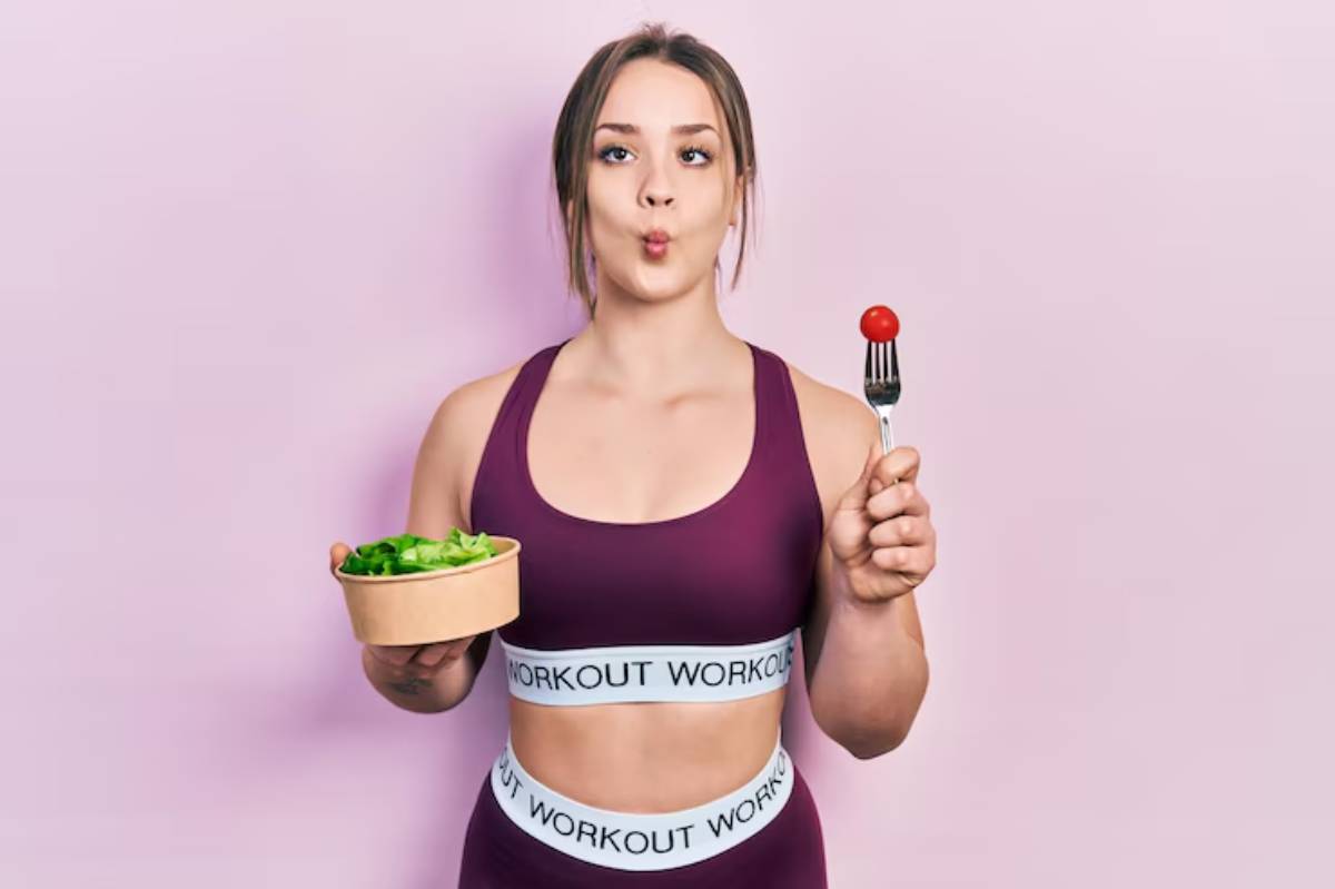 A person in athletic wear holds a bowl of salad in one hand and a fork with a cherry tomato in the other, set against a pink backdrop.