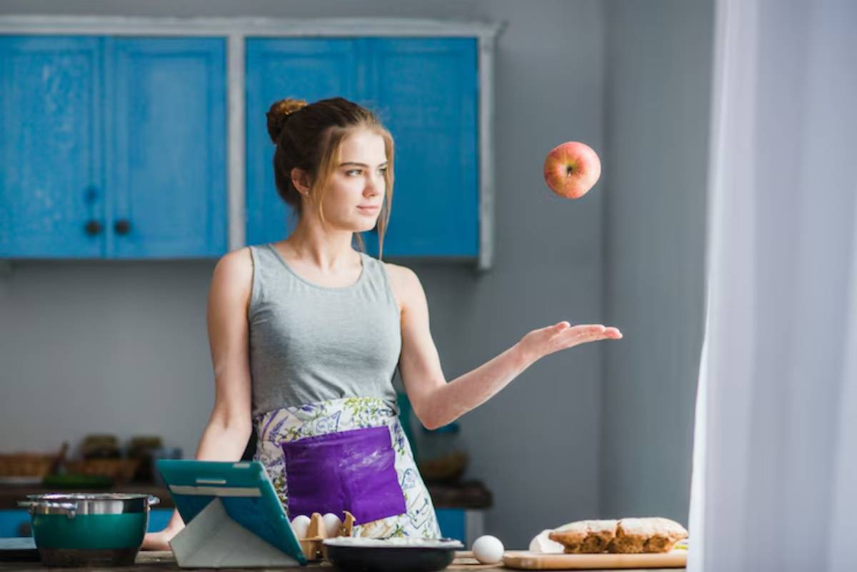 A person in a kitchen throws a red apple, surrounded by utensils, a pot, and baked goods on the counter, with blue cabinets in the background.