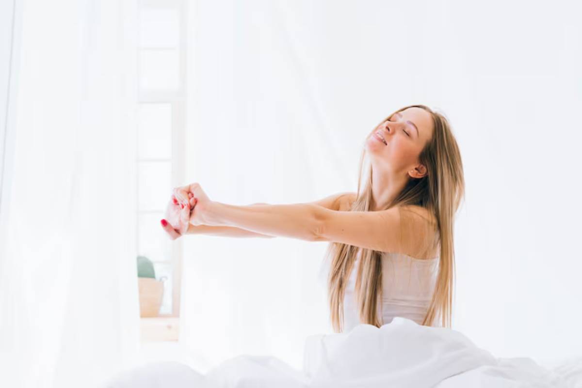 A woman stretches in bed, surrounded by soft white bedding and natural light streaming through sheer curtains.