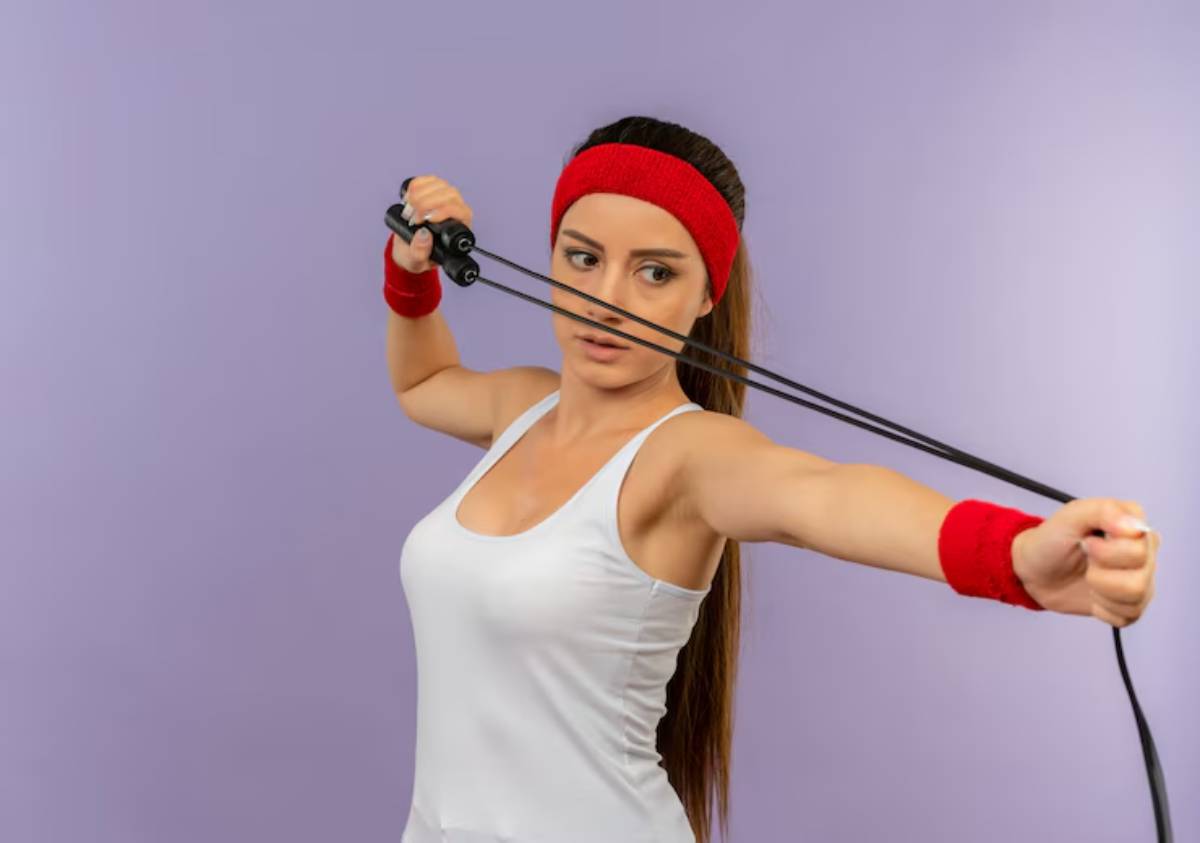 A person in a white tank top and red wristbands is stretching an exercise band against a light purple background.
