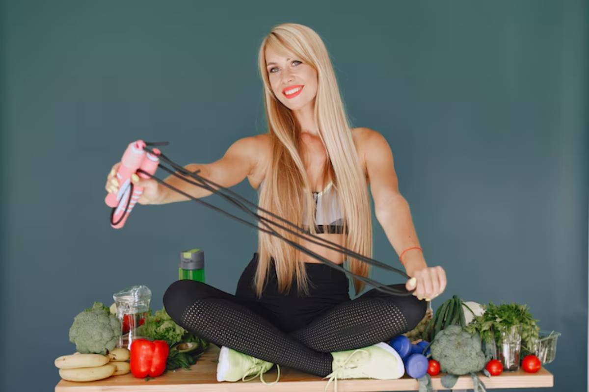 A fit individual sits cross-legged on a table surrounded by fresh vegetables and fruits, holding a resistance band for exercise.