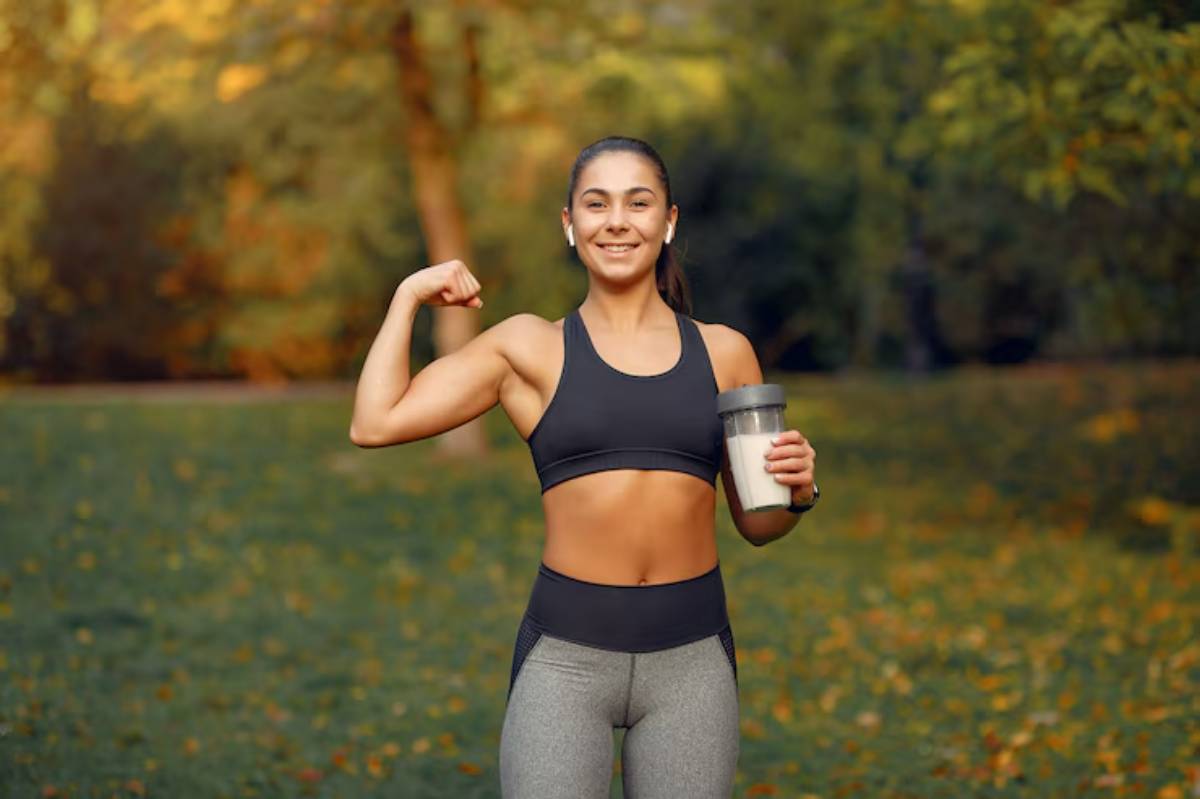 A confident woman in athletic wear flexes her bicep while holding a shaker bottle, surrounded by a lush, autumnal outdoor landscape.
