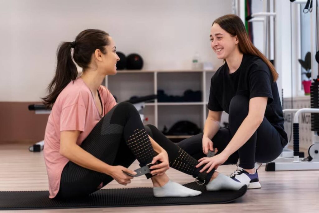 Two women are engaged in a fitness session, one adjusting straps on the other's legs while seated on a mat in a gym environment.