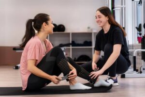 Two women are engaged in a fitness session, one adjusting straps on the other's legs while seated on a mat in a gym environment.