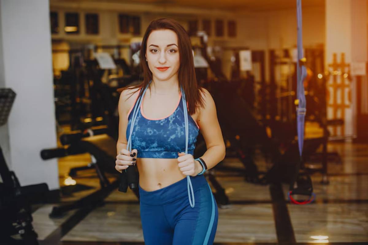 A fit woman in activewear holds a jump rope, jogging in a modern gym filled with workout equipment. Warm lighting enhances the scene.