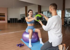 A woman balances on an exercise ball, engaging with a trainer who hands her a medicine ball in a modern gym setting.