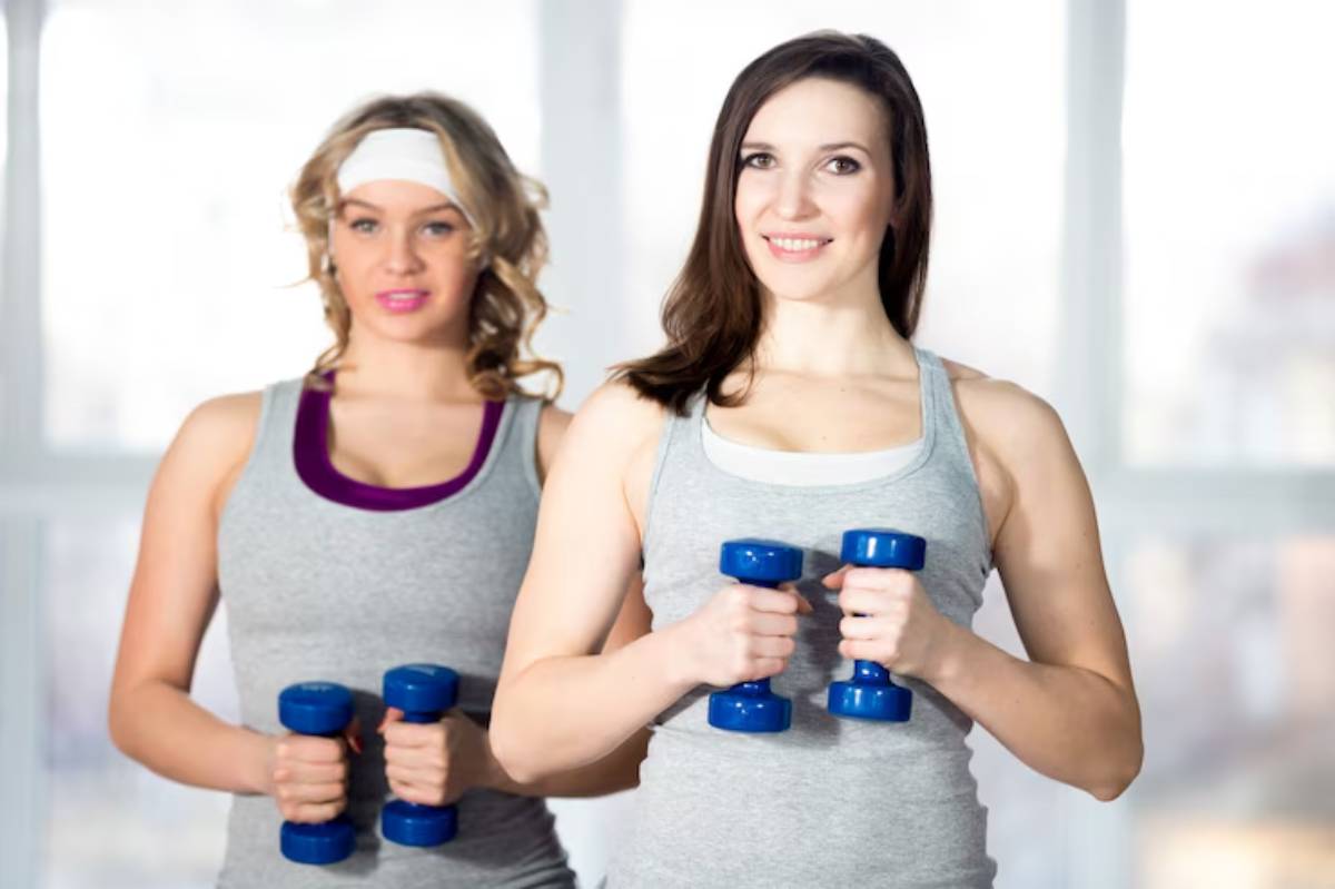 Two women in gray tank tops hold blue dumbbells in a bright, well-lit gym setting, ready for a workout.