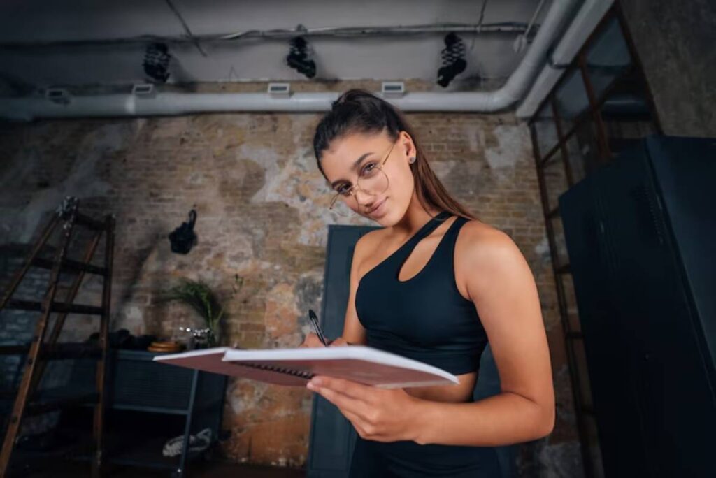A woman in a black athletic outfit holds a notebook and pen, standing in a stylish, industrial-style room with exposed brick walls.