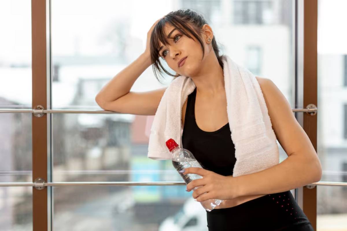 A person in athletic attire, holding a water bottle, leans against a window with a towel draped around their neck, looking tired after a workout.