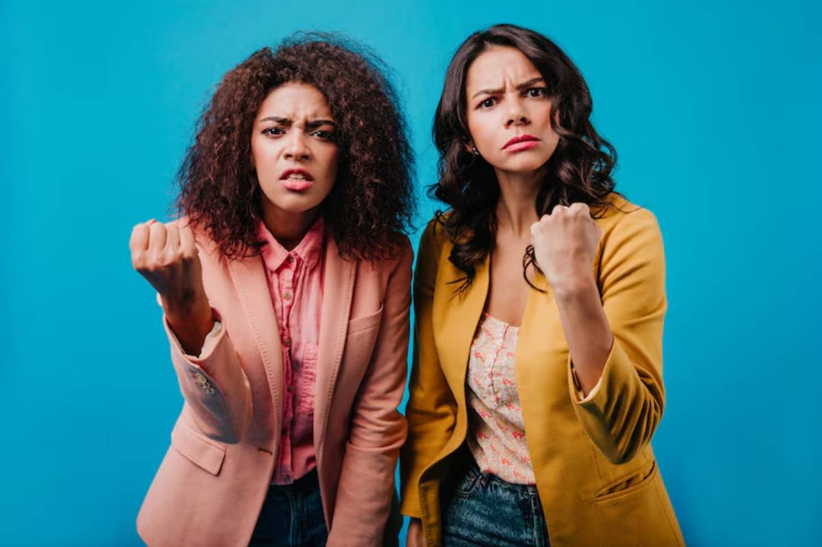 Two women strike confident poses against a bright blue background, wearing stylish blazers and showcasing empowering fist gestures.