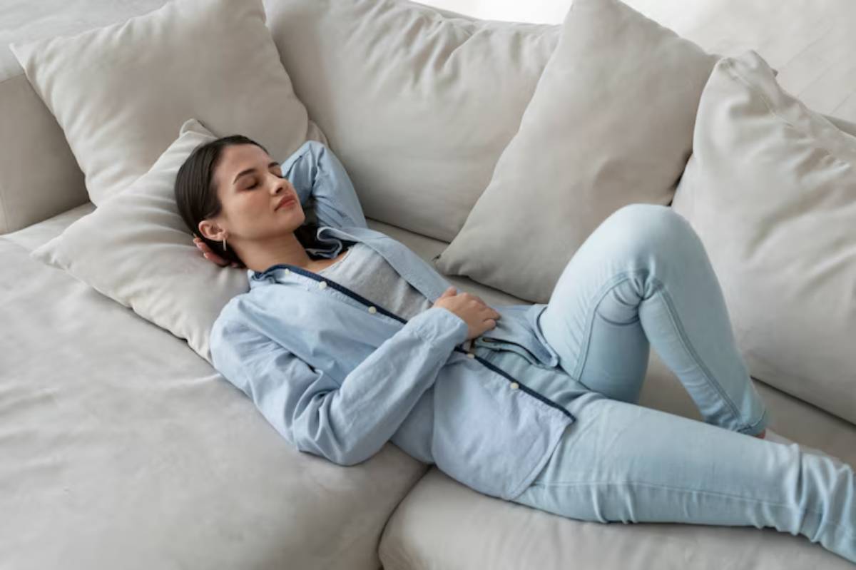 A person relaxes on a light-colored sofa, wearing a blue shirt and light jeans, with pillows surrounding them.