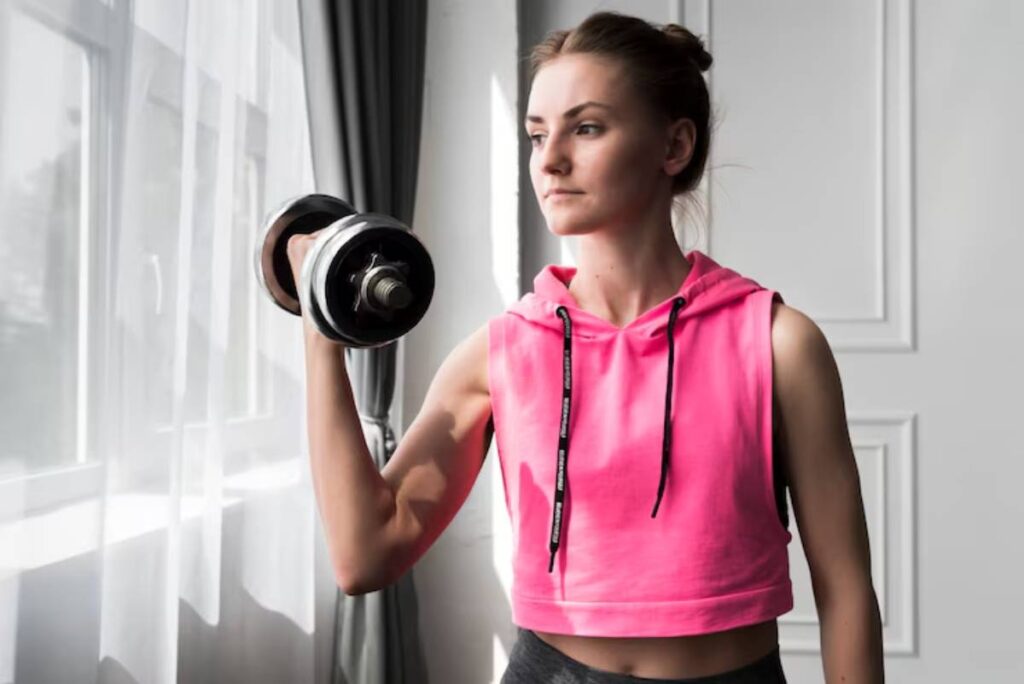 A person in a pink cropped hoodie lifts a dumbbell, showcasing a strong arm, with sunlight streaming through a window.