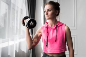 A person in a pink cropped hoodie lifts a dumbbell, showcasing a strong arm, with sunlight streaming through a window.
