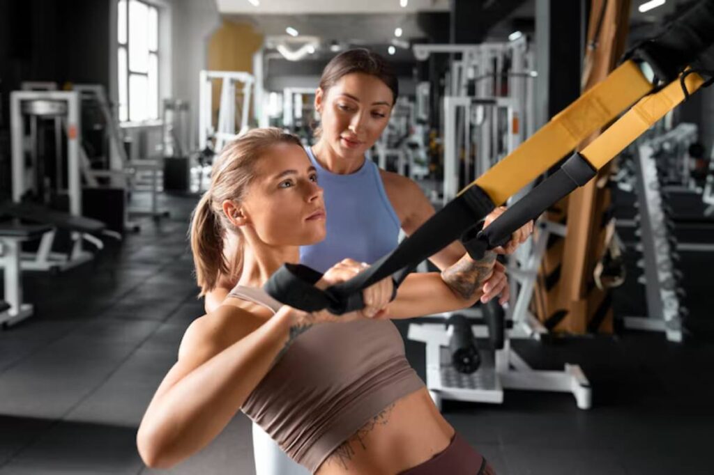 A woman engages in a TRX workout, receiving guidance from a trainer in a modern gym setting filled with fitness equipment.
