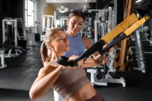 A woman engages in a TRX workout, receiving guidance from a trainer in a modern gym setting filled with fitness equipment.