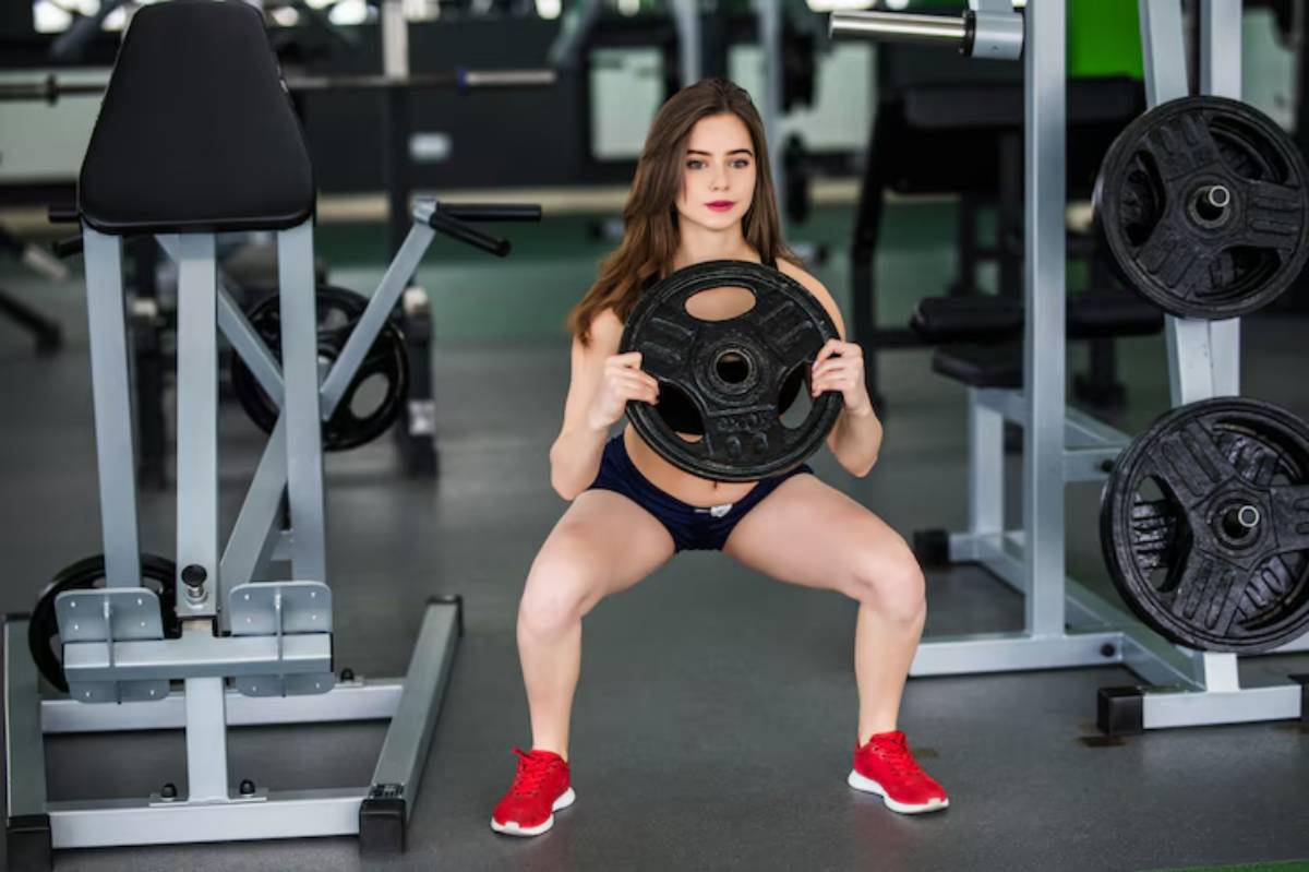 A person squats while holding a weight plate in a gym, surrounded by exercise equipment and weights. They're focused on their workout.