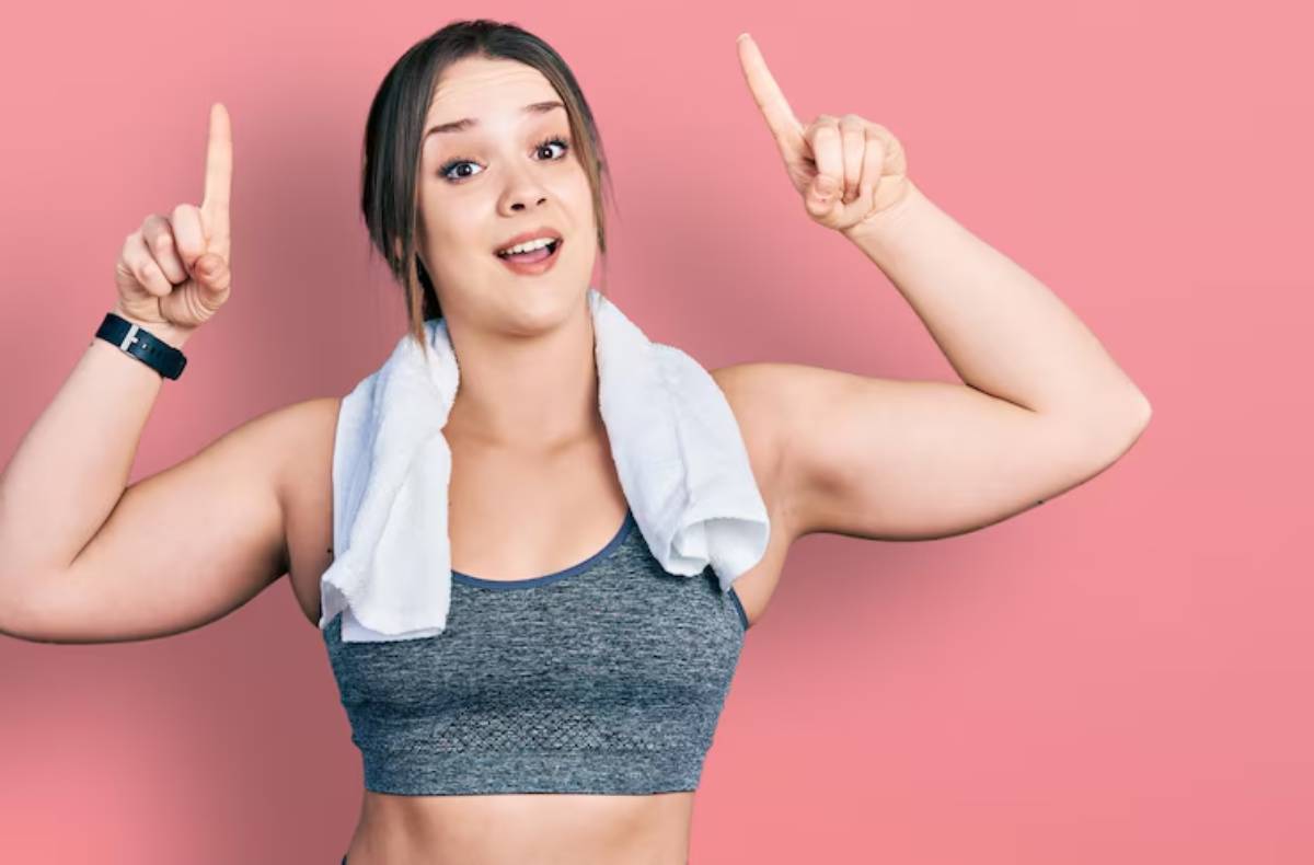 A woman in a gray sports bra and white towel raises both hands, smiling, against a pink background, showcasing empowerment and fitness.