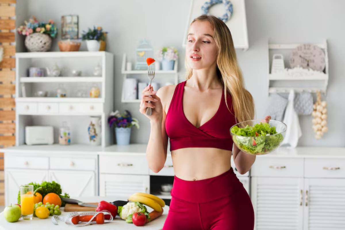 A woman in a red workout outfit holds a bowl of salad and a fork, surrounded by fresh fruits and vegetables in a bright kitchen.
