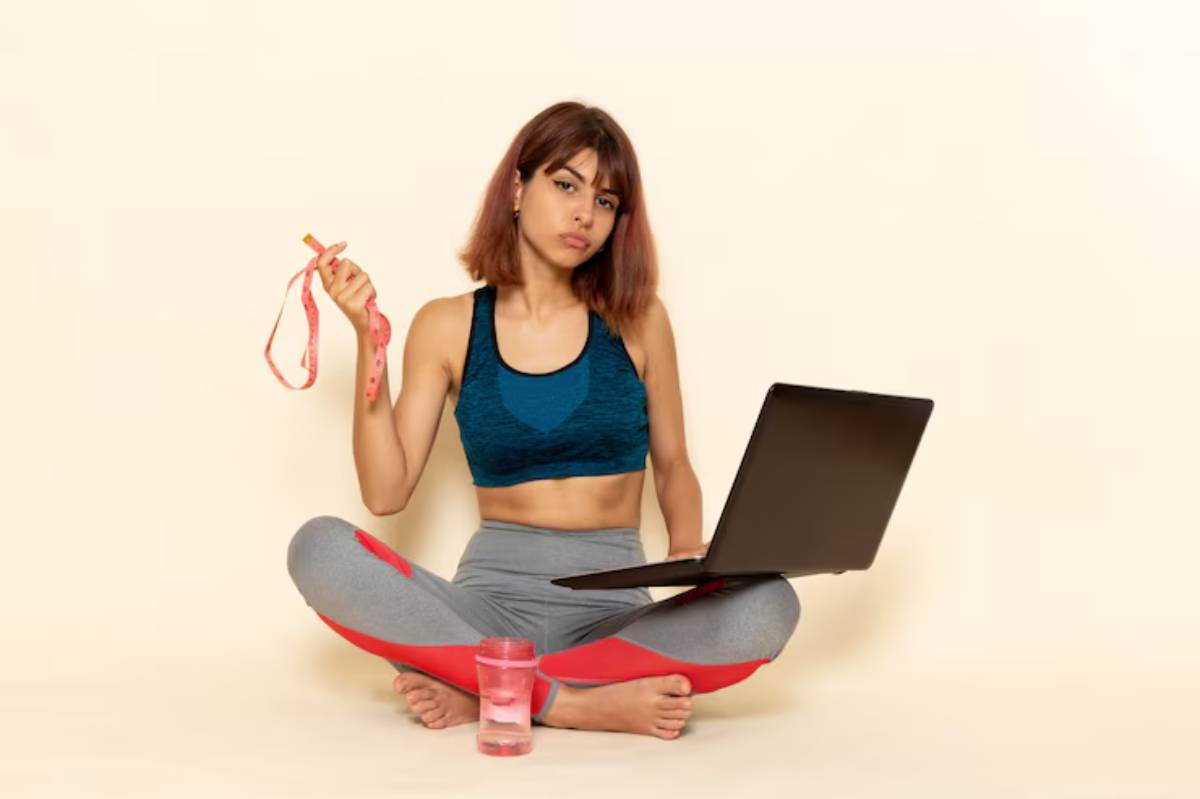 A person sits cross-legged on the floor, holding a tape measure in one hand and a laptop in front, with a glass of water beside them.
