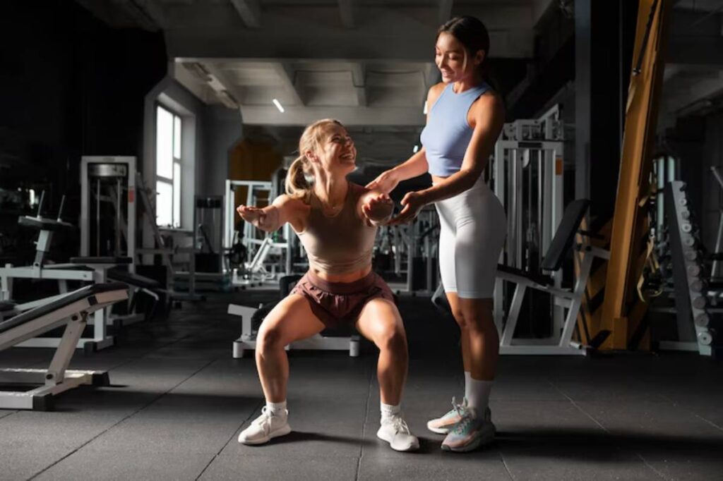A woman is performing squats in a gym, supported by a trainer. Various gym equipment is visible in the background.
