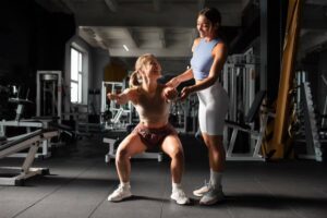 A woman is performing squats in a gym, supported by a trainer. Various gym equipment is visible in the background.
