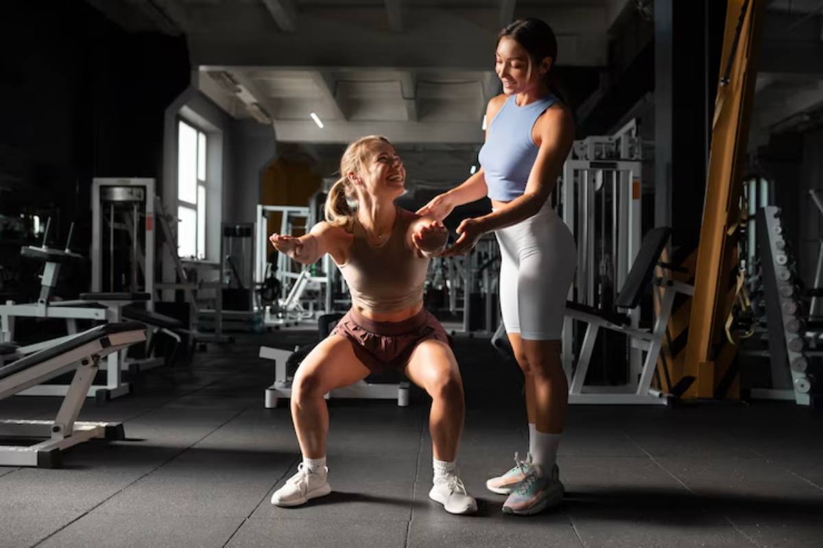 A woman is performing squats in a gym, supported by a trainer. Various gym equipment is visible in the background.