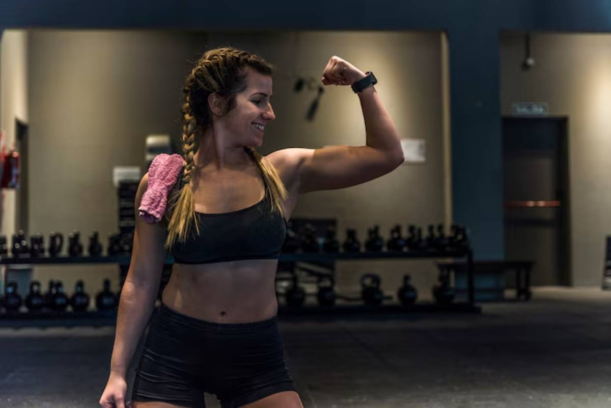 A fit woman flexes her bicep in a gym, wearing a black sports outfit and a towel draped over her shoulder, with weights in the background.
