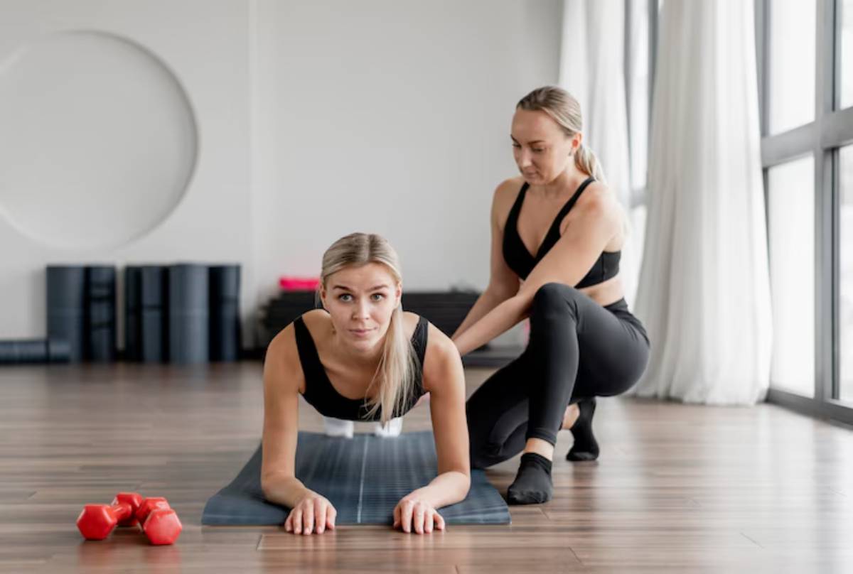 A fitness instructor assists a woman in a plank position on a yoga mat, with dumbbells nearby and large windows letting in natural light.
