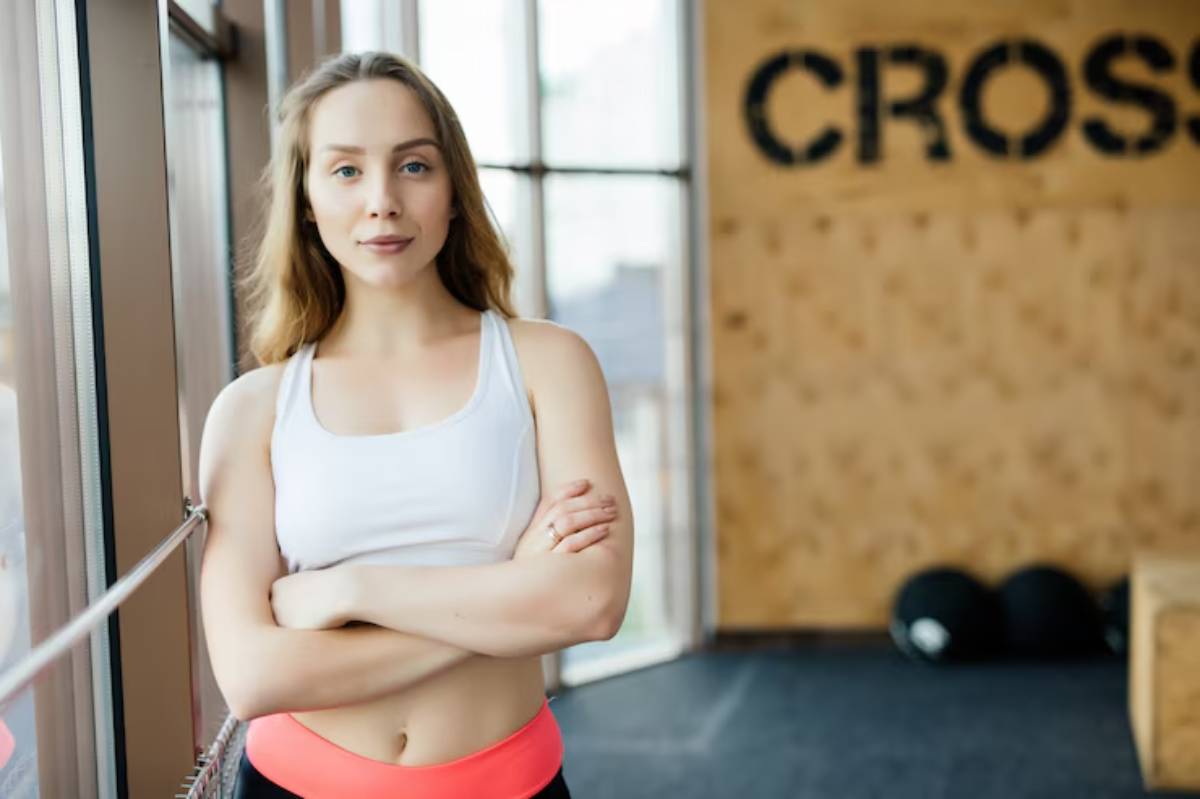 A fit woman in a gym wearing a white tank top and bright pink leggings stands confidently with arms crossed, overlooking the space.