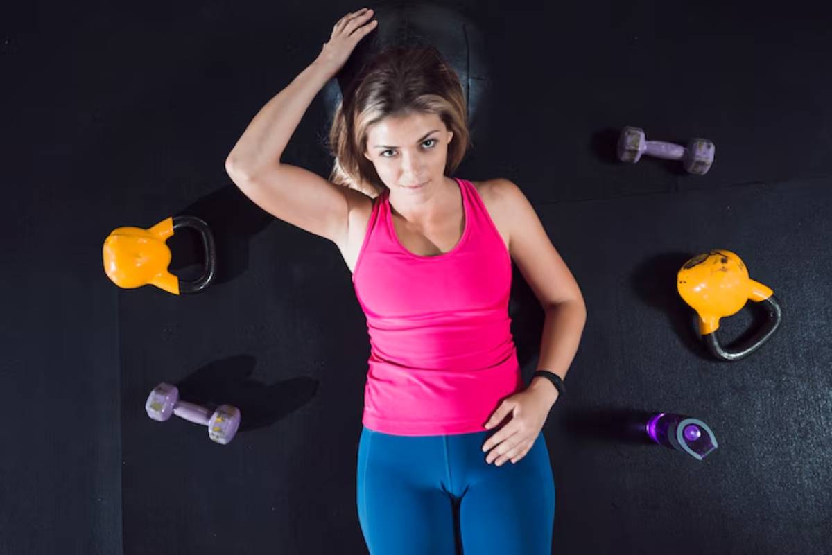 A woman in a pink workout tank top and blue leggings lies on a gym floor surrounded by kettlebells and dumbbells.