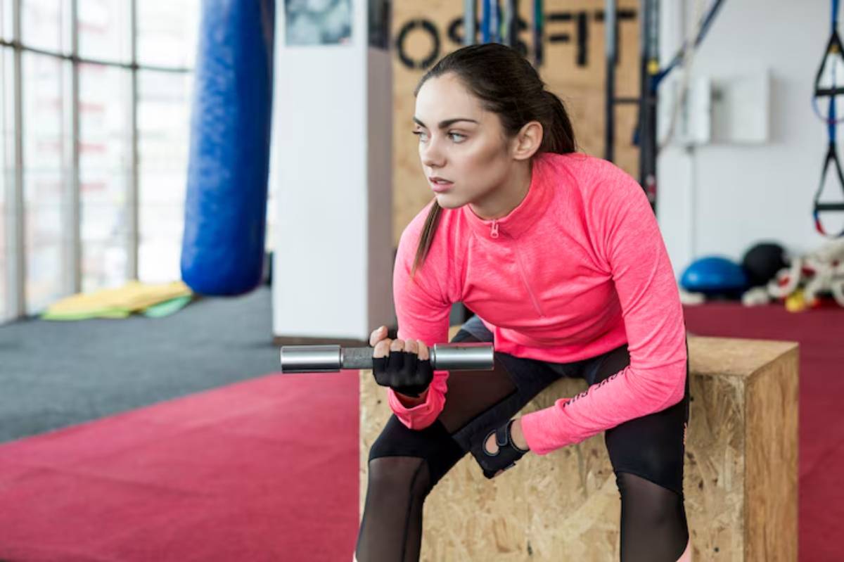 A person in a bright pink athletic top and gloves sits on a wooden box, holding a barbell in a gym setting.