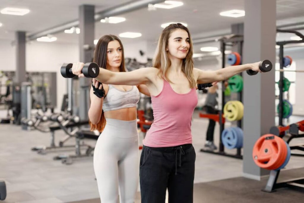 A woman in a pink tank top performs a shoulder exercise with dumbbells, while another person supports her in a modern gym setting.