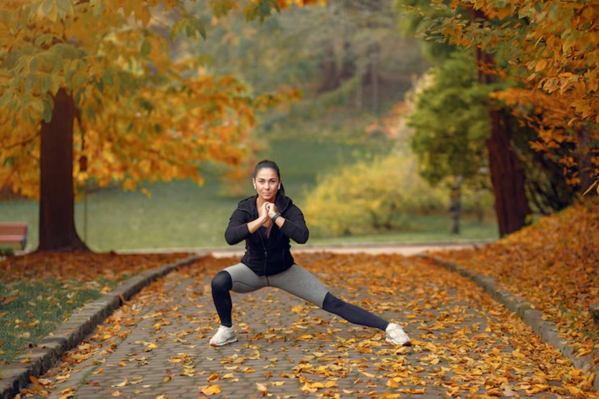 A person in athletic wear performs a squat on a stone path covered with autumn leaves, surrounded by trees with colorful foliage.