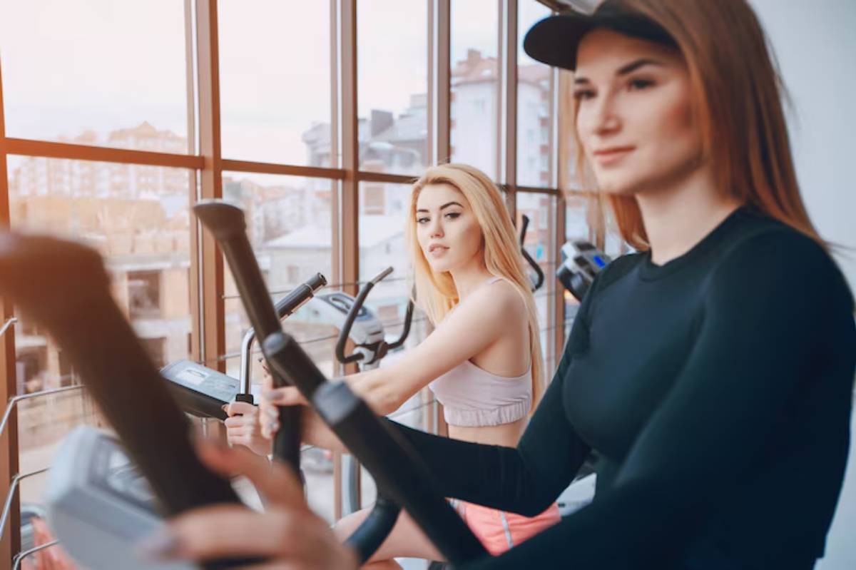 Two women exercise on elliptical machines in a bright gym with large windows showcasing a city view.