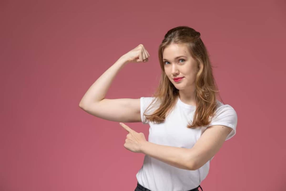 A woman in a white t-shirt flexes her bicep and points with her other hand, set against a pink background.