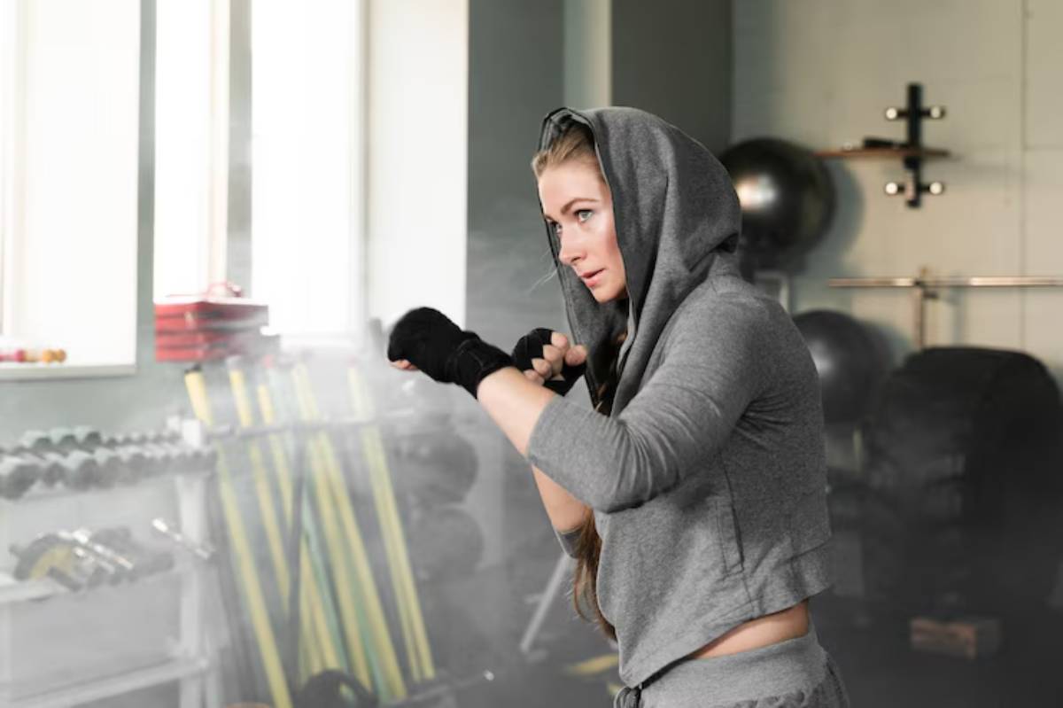 A person in a gray hoodie practices boxing in a gym, surrounded by weights and exercise equipment in a well-lit space.