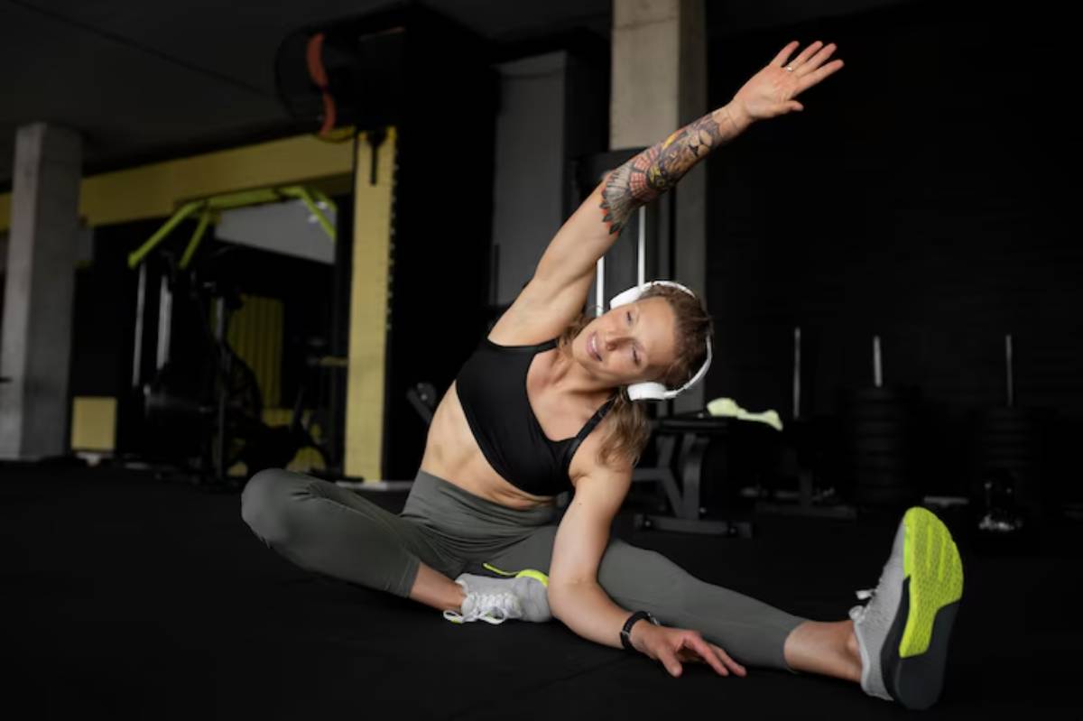 A fit individual stretches in a gym, wearing a black top, gray leggings, and sneakers, showcasing a tattooed arm in an athletic pose.