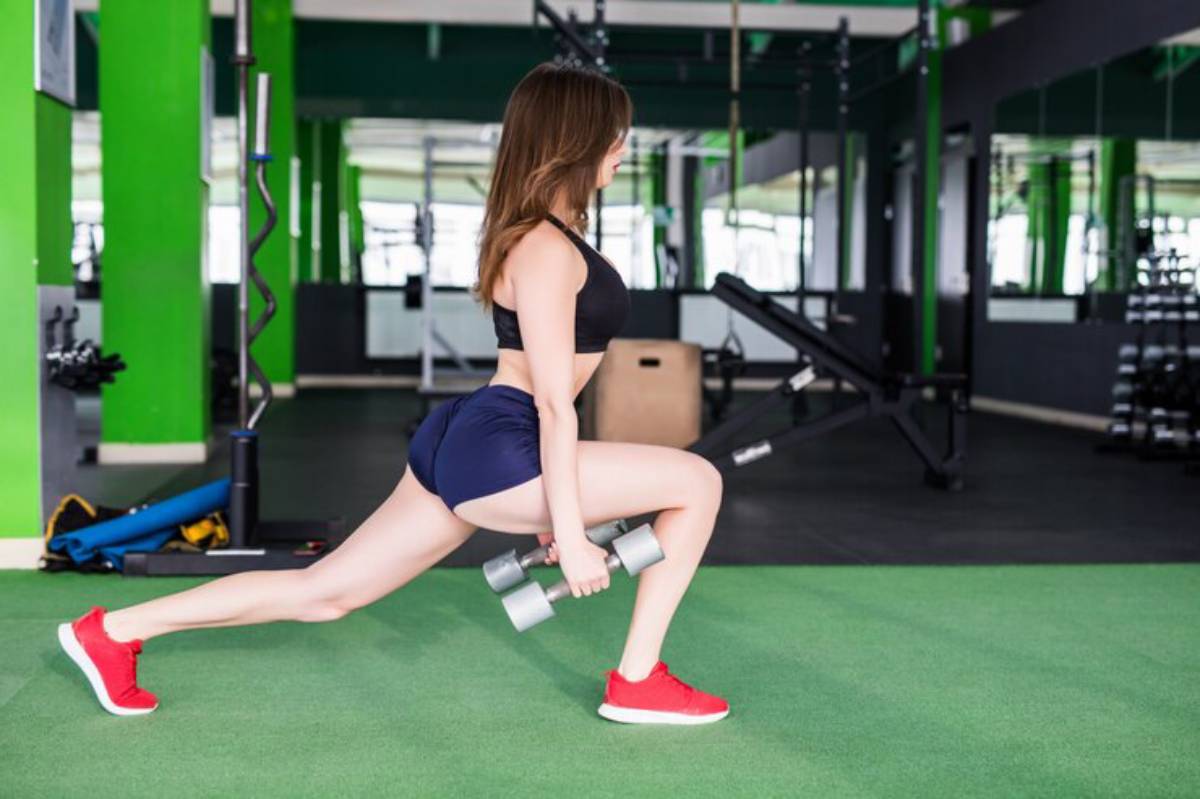 A young woman performs a weighted lunge in a gym, wearing a black sports bra and shorts, with green walls and equipment in the background.