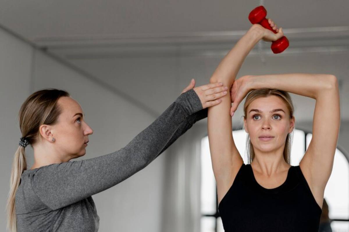 A woman in a black tank top lifts a red dumbbell overhead while another woman adjusts her arm in a bright training space.