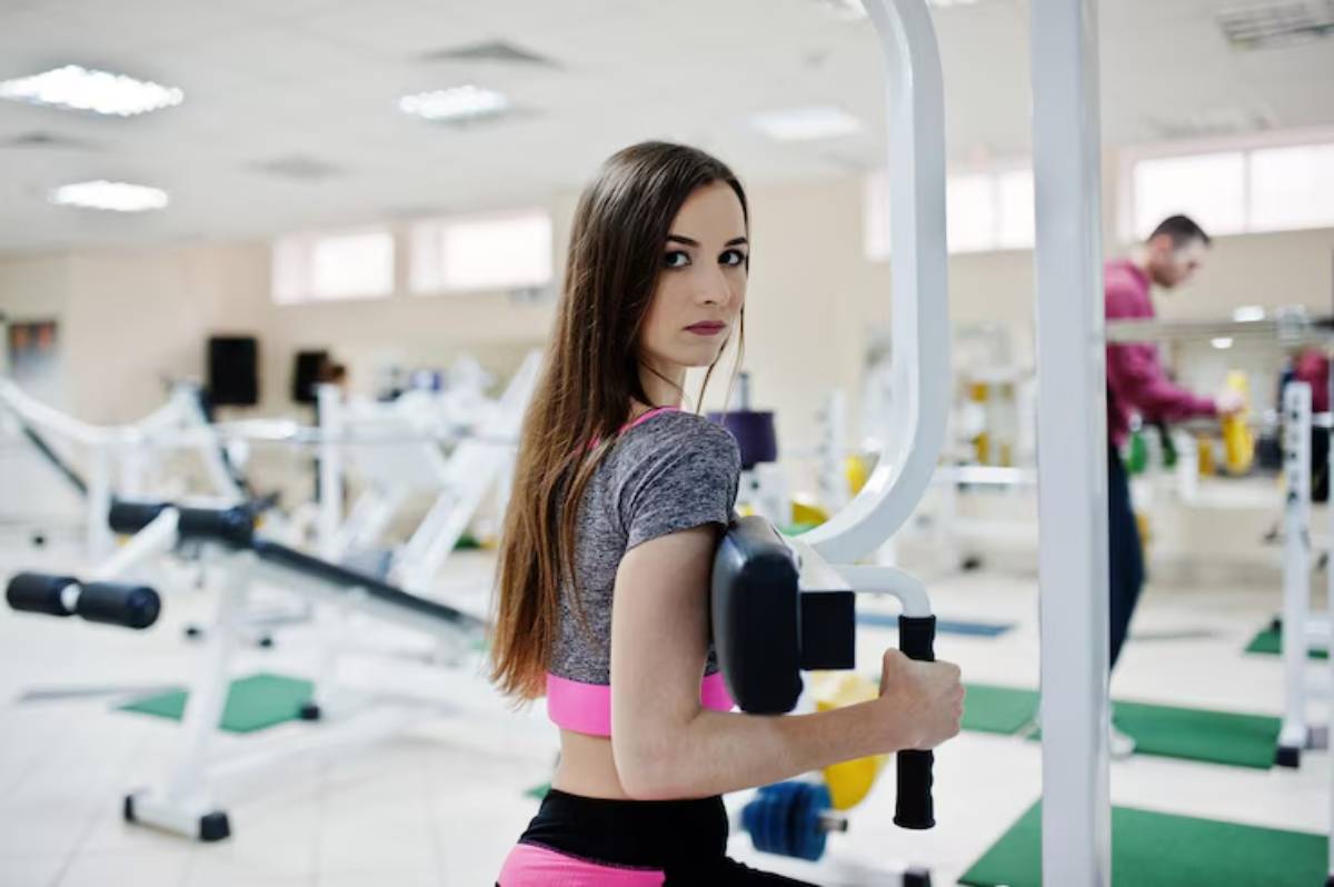 A young woman exercises in a gym, using a weight machine while wearing a gray crop top and pink workout gear. Equipment is visible in the background.