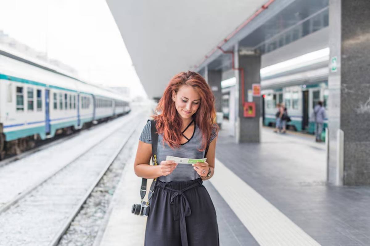 A young woman stands at a train station, holding a ticket and wearing a striped shirt and black pants, with a camera slung over her shoulder.