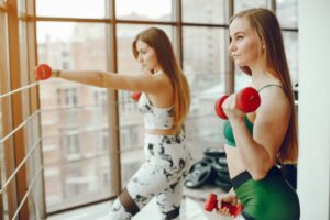 Two women with long hair workout at a gym, one lifting dumbbells while the other performs an exercise against a railing by large windows.