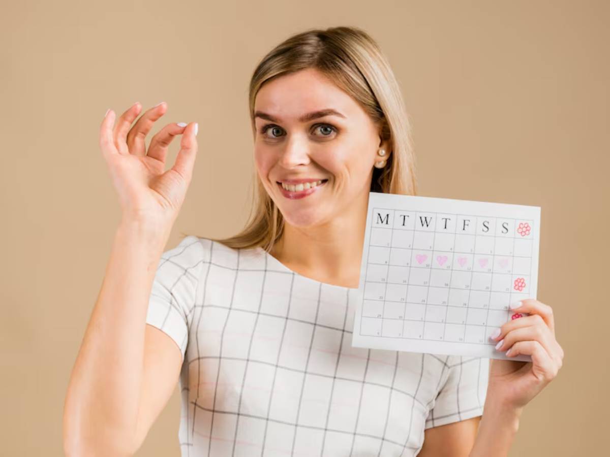 A woman in a grid-patterned shirt holds a calendar with marked dates, smiling and gesturing with her other hand.