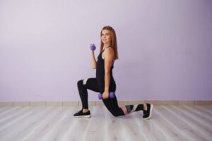 A person is performing a lunge with a dumbbell, wearing workout attire, against a light purple wall. The floor is light-colored wood.
